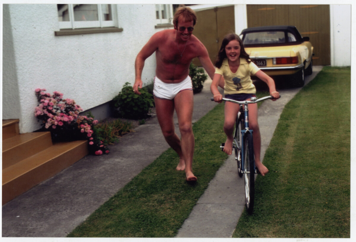 Colour photo of a girl riding a bike in a driveway with a man pushing her